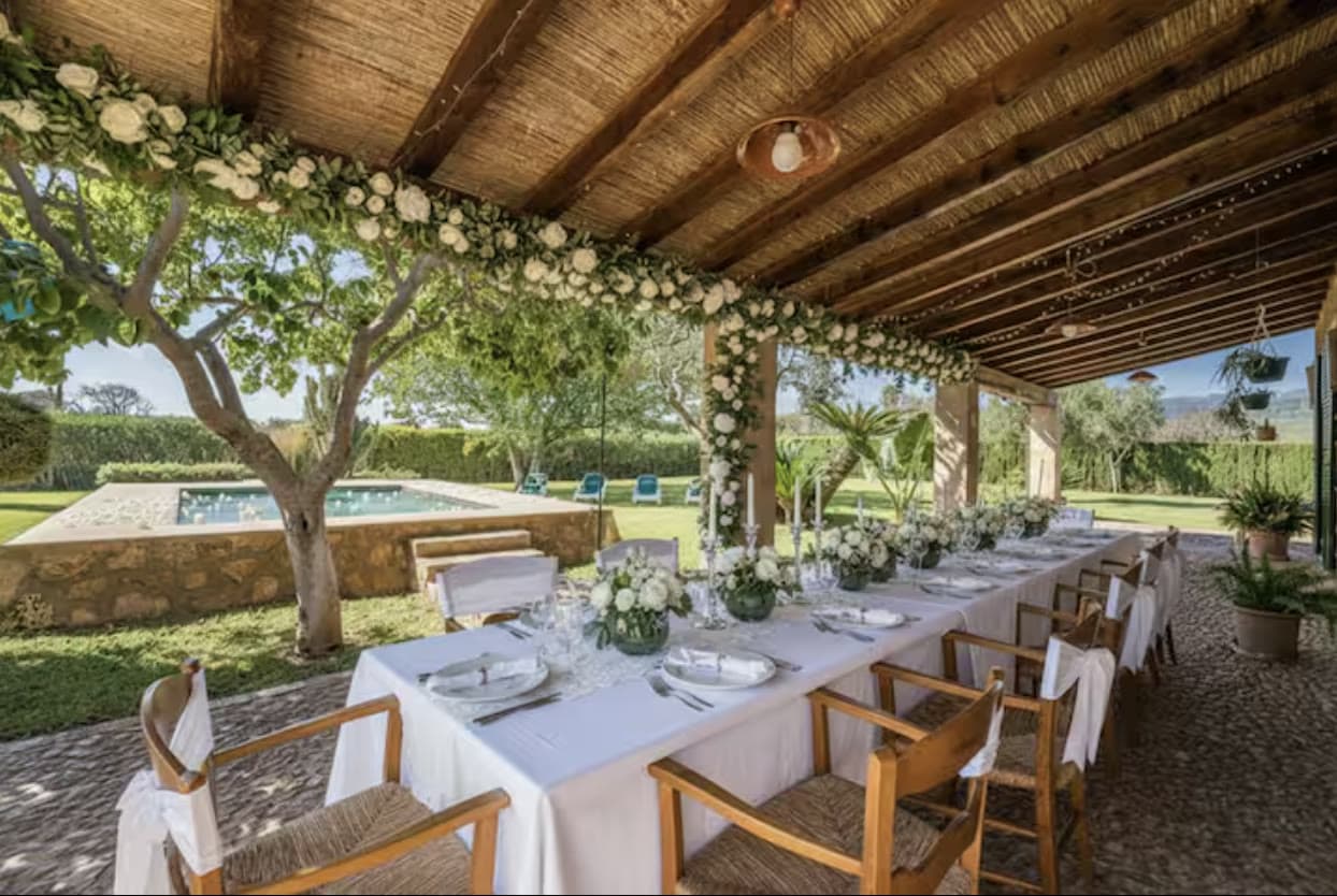 A long dinner table set with white flowers under the finca's covered terrace, beside the pool